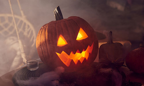 A pumpkin with a scary face cut out of it with a light inside surrounded by smoke, cobwebs, and candles